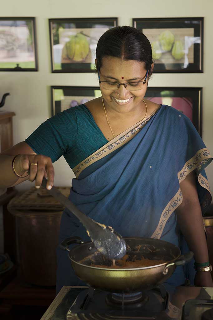 Administrator and chef's assistant Onamly, wearing a jewel toned blue sari with gold trim, smiles as she gently stirs food cooking simmering in a wok.