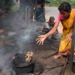 A young woman in a yellow and orange sari with a pink top is in a bent position while tossing cashews with an extended right arm into an old iron pot placed on a fire on the dirt ground.