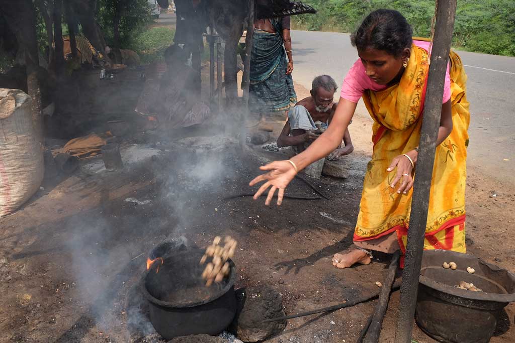 A young woman in a yellow and orange sari with a pink top is in a bent position while tossing cashews with an extended right arm into an old iron pot placed on a fire on the dirt ground.