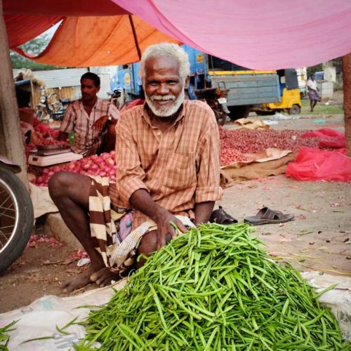 A smiling man in an orange checked button up shirt and striped dhoti sells long green beans in a vegetable market outside of Madurai.