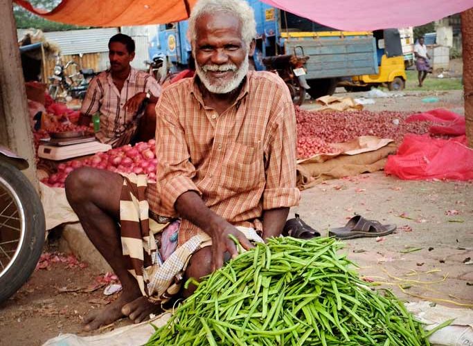 A smiling man in an orange checked button up shirt and striped dhoti sells long green beans in a vegetable market outside of Madurai.