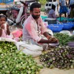 A man selling vegetables sits on a tarp amongst his large piles of eggplant. He passes a customer a handful of purple eggplant while a young girl in a pink dress and pig tales drinks from a small white cup and looks at the camera.