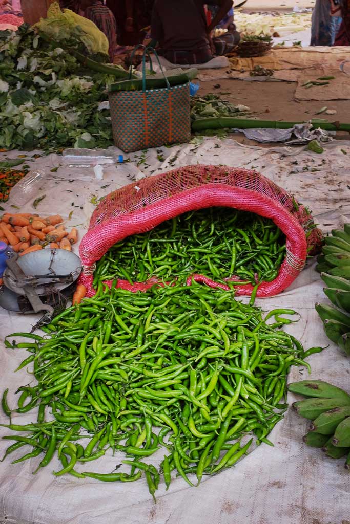 Long green chillies spill out of a red bag sitting open on white tarp laid on the ground in a vegetable market outside of Madurai.