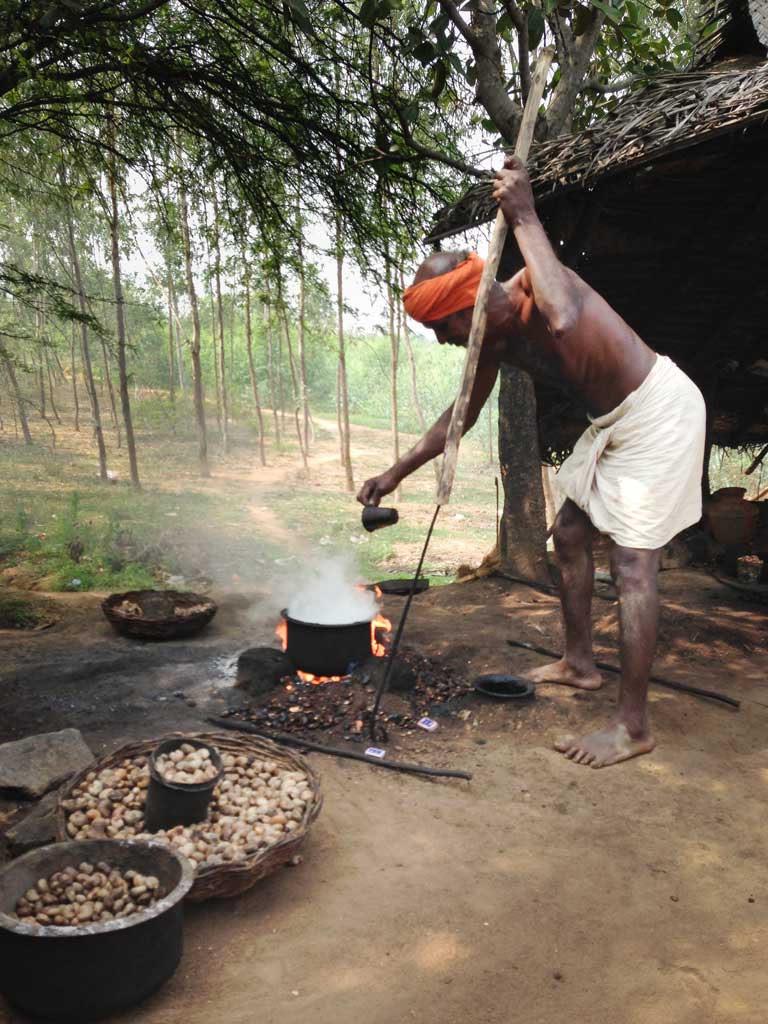 A shirtless South Indian man wearing an orange head wrap and a white dhoti carries a long wooden pole with a metal poker end. He leans forward against the pole as he pours a cup of water into his steaming pot, which sits on a ground level fire. One stone bowl and one basket of roasted cashews sit in the foreground.