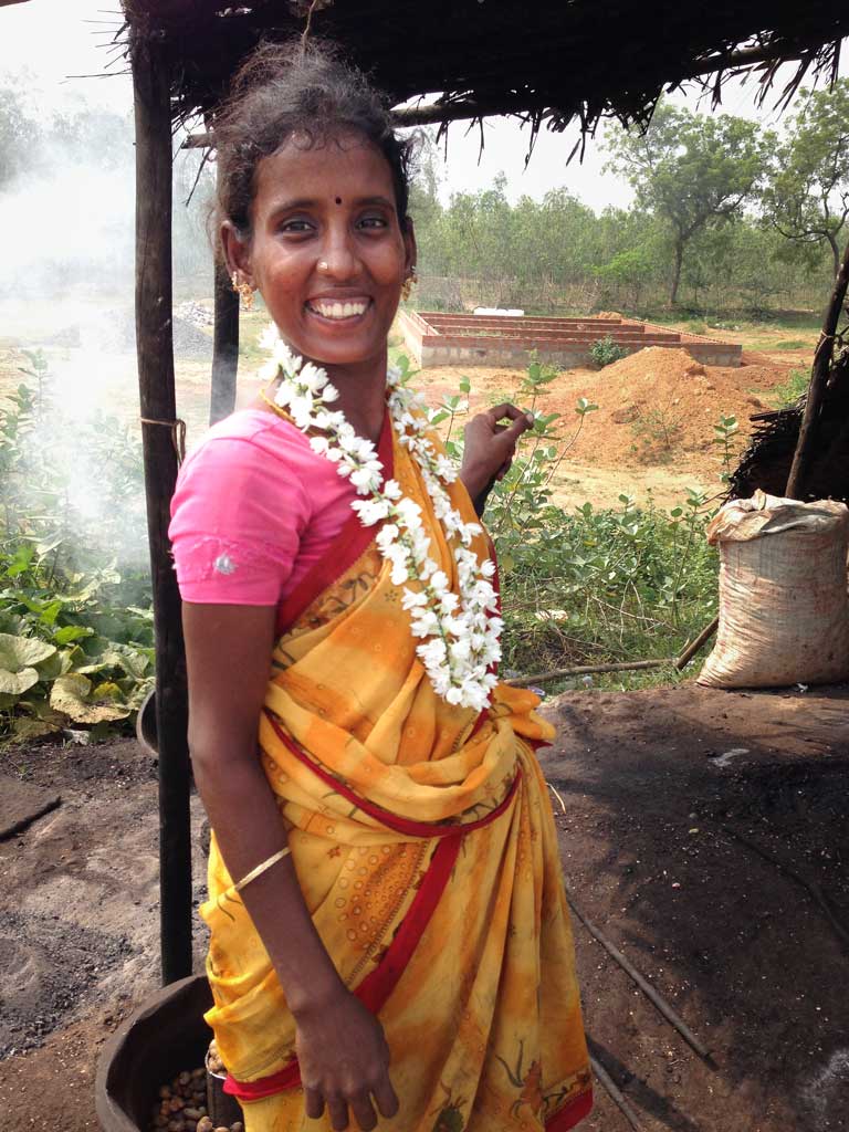 A young South Indian woman wearing a yellow and orange sari with a pink top and a necklace of jasmine smiles at the camera. Smoke from her roasting cashews rises behind her.