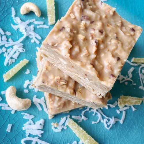 Three 2" squares of creamy, nutty cashew coconut fudge sit on a turquoise blue plate stacked in a pile one on top of the other, with garnishes of white shredded coconut, yellow candied ginger strips and raw cashews strewn about on the plate.