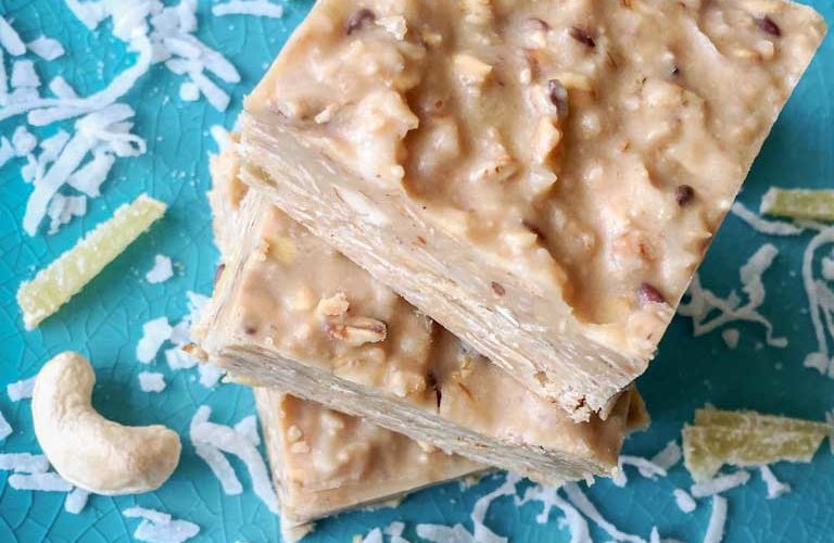 Three 2" squares of creamy, nutty cashew coconut fudge sit on a turquoise blue plate stacked in a pile one on top of the other, with garnishes of white shredded coconut, yellow candied ginger strips and raw cashews strewn about on the plate.