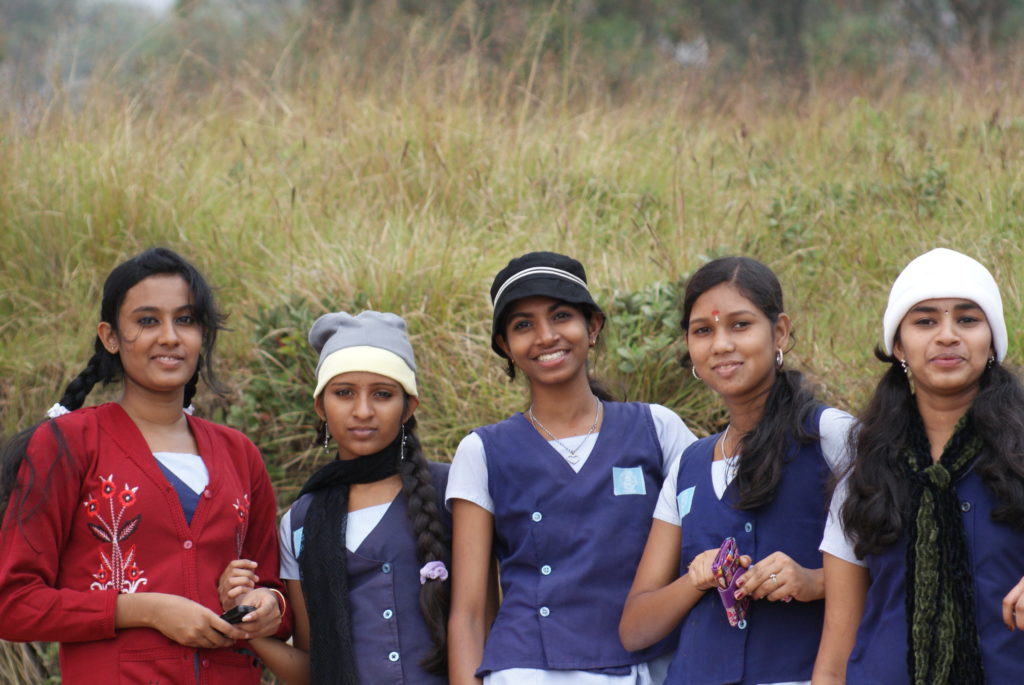 Five South Indian school girls dressed in purple uniforms pose and smile in front of a field of grass.