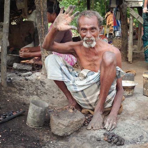 A shirtless, elderly South Indian man wearing a light coloured dhoti squats on the ground with his knees near his shoulders as he waves open-palmed to the camera. Unshelled roasted cashews and a grinding stone sit at his feet.