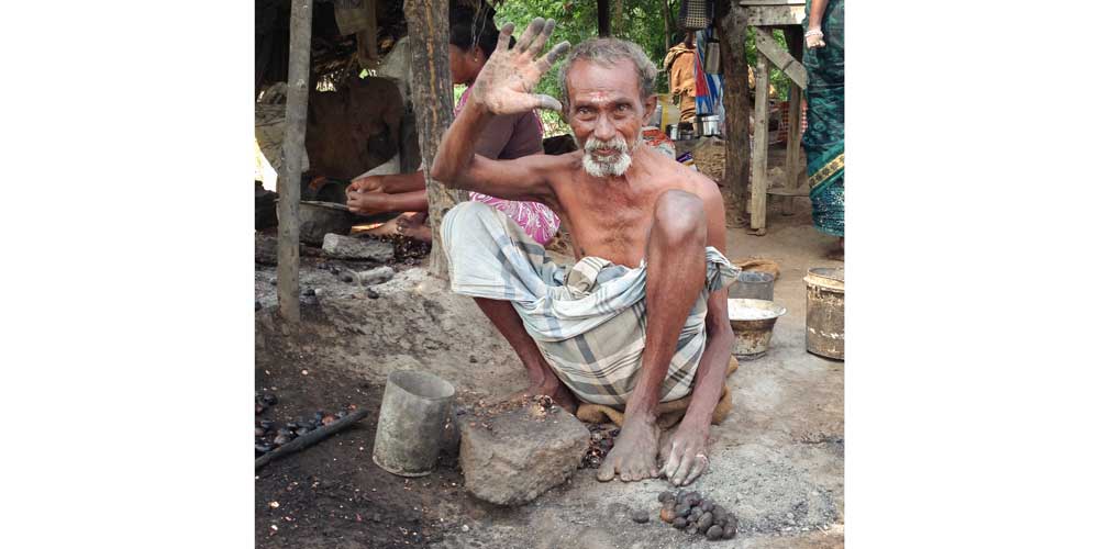 A shirtless, elderly South Indian man wearing a light coloured dhoti squats on the ground with his knees near his shoulders as he waves open-palmed to the camera. Unshelled roasted cashews and a grinding stone sit at his feet.