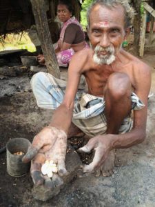 A shirtless, elderly South Indian man wearing a light coloured dhoti squats on the ground with his knees near his shoulders as he extends his hand out to the camera and offers a palm full of freshly roasted cashew nuts.