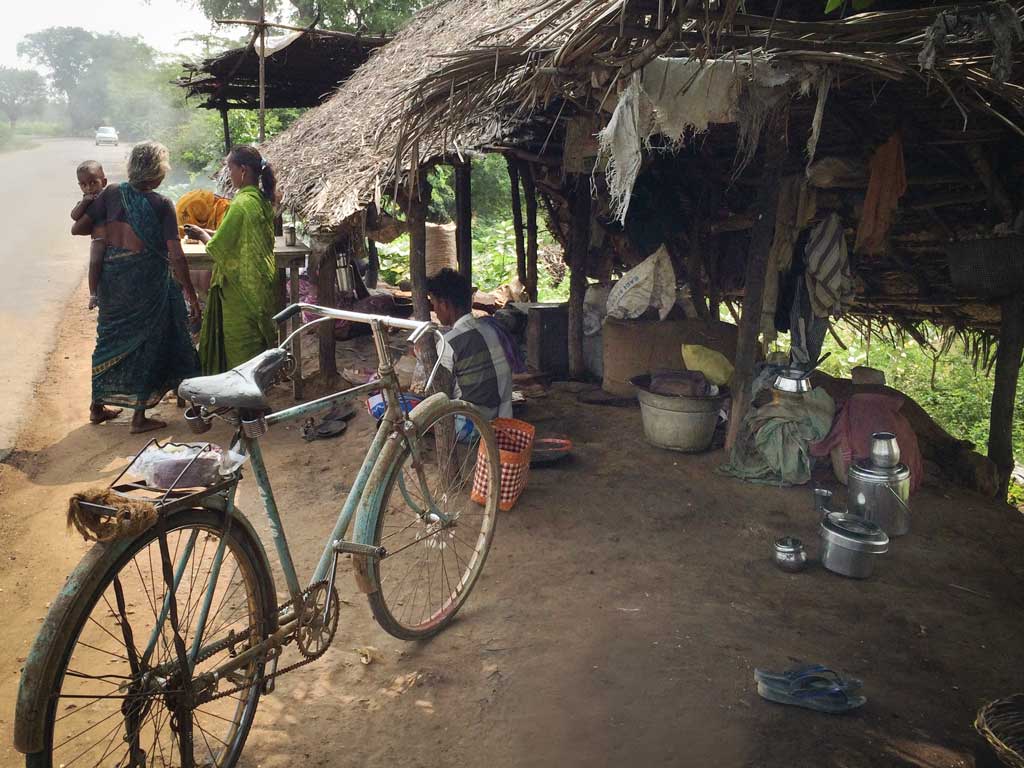 An old bicycle waits in the foreground of a thatched roofed, open-air cashew roasting operation.