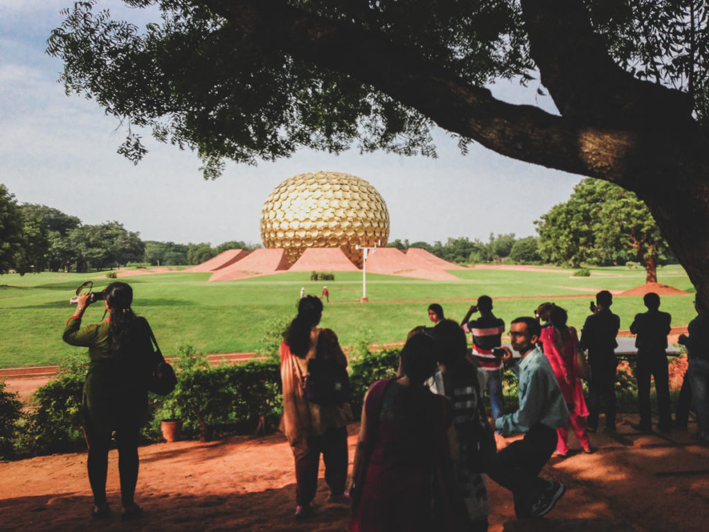 A crowd gathers under the shade of trees to observe the Matrimandir in Auroville.