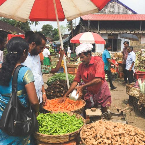 A man sells vegetables under a red and white umbrella at the Kalpaka vegetable market.