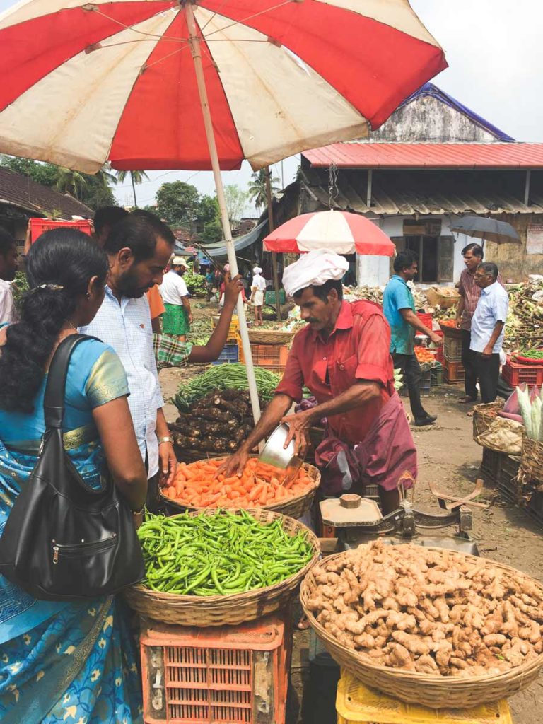 A man sells vegetables under a red and white umbrella at the Kalpaka vegetable market.