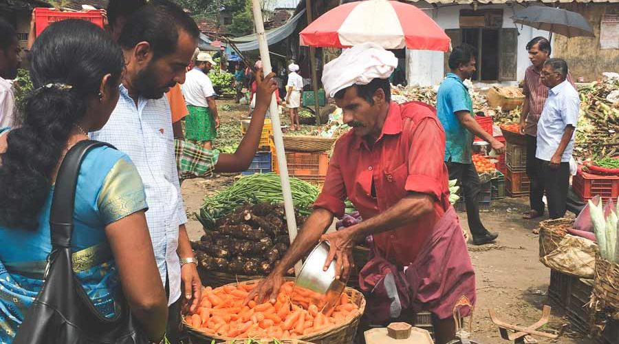 A man sells vegetables under a red and white umbrella at the Kalpaka vegetable market.