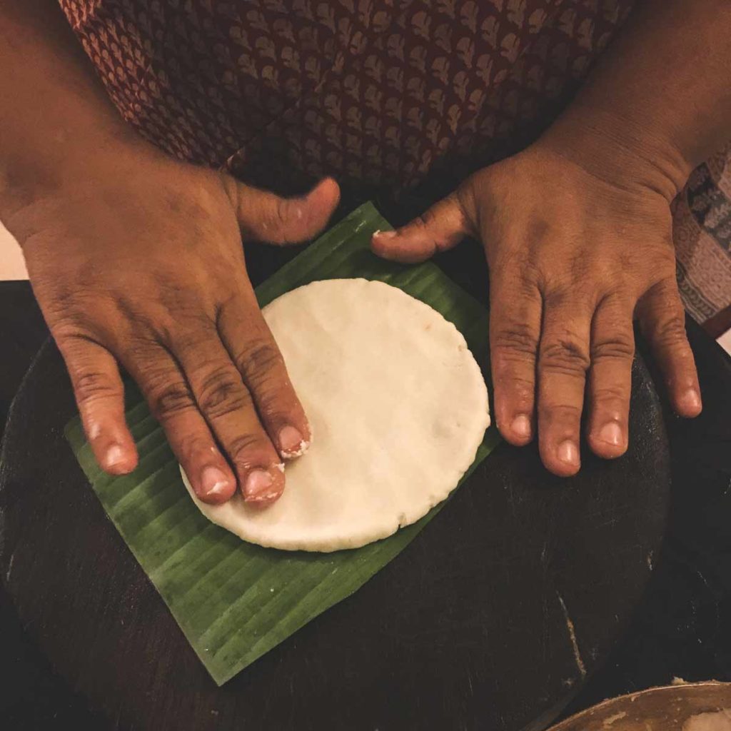 A hand pats flat Pathiri bread dough on a banana leaf.